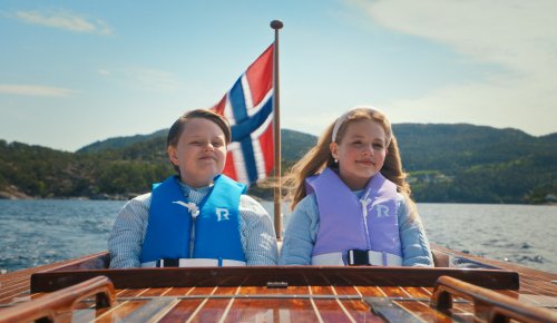 foto: en gutt og en jente sitter baki en båt med redningsvester foran et norsk flagg i norsk fjordlandskap