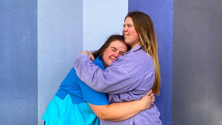 foto. to kvinner i halvfigur foran  en vegg i forskjellige blåfarger står og holder rundt hverandre