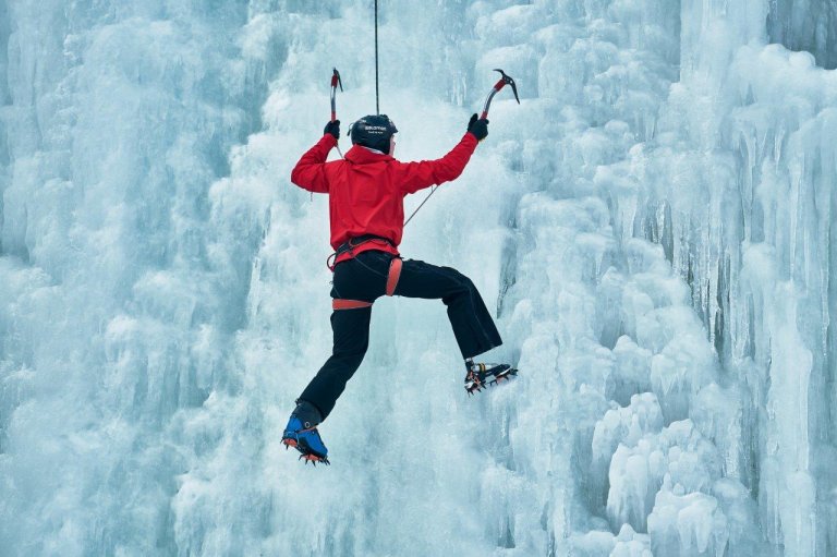 foto. en fjellklatrer kledt i rød jakke henger i en isvegg, hans høyre fot er en protese
